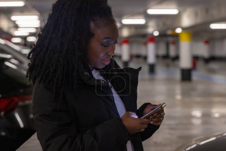 The image portrays a young woman engaged with her smartphone in a dimly lit parking garage. Her long, black dreadlocks cascade over her shoulders, and she is dressed in a casual black jacket, which