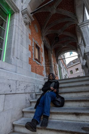 This image captures a young woman in a moment of repose on the steps of a historic building. The architectural details, including the arched passageway and brickwork, provide a classic European feel