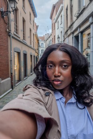 A young African woman with dark hair is taking a selfie in a quaint European alleyway. She is dressed casually in a blue striped shirt and a beige trench coat. Her expression is confident and direct