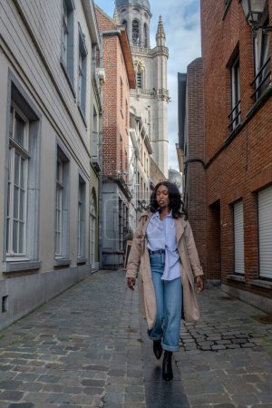 The image captures a poised African woman enjoying a leisurely walk along a cobblestone lane in a historical European city. She is fashionably dressed in a casual chic ensemble consisting of a light