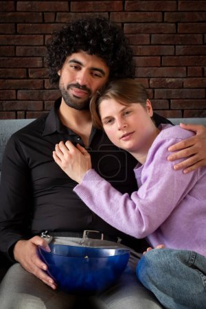 A couple enjoys a relaxing moment on the couch in a homely atmosphere. The man, with curly black hair and a beard, exudes a sense of contentment as he holds a remote, suggesting they are settled in