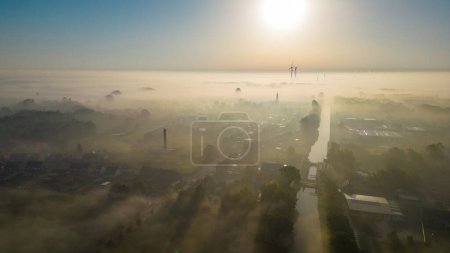This aerial image captures an urban landscape veiled in morning fog at dawn, creating an ethereal effect. The sun, low on the horizon, bathes the city in a soft, golden light. The fog adds a layer of