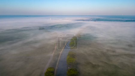 Esta fotografía aérea captura la tranquila belleza de un canal que atraviesa un paisaje envuelto en niebla al amanecer. La suave luz de la madrugada se difunde suavemente a través de la niebla