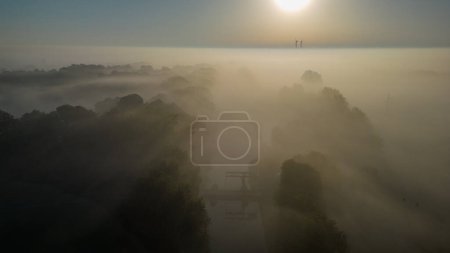 A serene misty morning over an urban park is beautifully captured in this aerial photograph. The rising sun casts a soft, diffuse glow, gently highlighting the tops of the trees and buildings cloaked