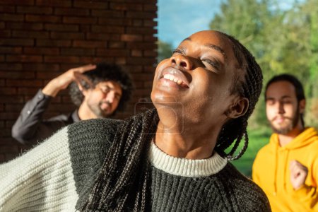 In einem Raum, der in das warme Licht der Sonne getaucht ist, verkörpert eine afroamerikanische Frau das Bild des Überschwangs, den Kopf vor Lachen zurückgeworfen, einen Moment reiner Freude und Befreiung verkörpernd. Ihre geflochtenen