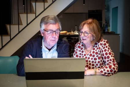 The image depicts an elderly couple jointly focused on a laptop screen. The man, wearing glasses and a navy jacket over a blue shirt, is intently observing the screen. Next to him, a woman in a polka