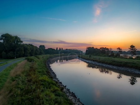 Cette image tranquille capte la première lumière de l'aube se reflétant sur les eaux calmes d'un canal, avec les teintes pastel du ciel matinal reflétées sur sa surface. Un sentier longe le canal