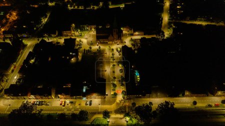 This nighttime aerial image captures the ambient illumination of a town square, where the warm glow of street lights creates a network of visibility against the dark backdrop of the night. The central