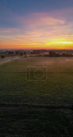 Captured at the break of dawn, this image encapsulates the serene beauty of a countryside scene bathed in the soft, warm glow of sunrise. The horizon is painted with a palette of rich oranges, pinks