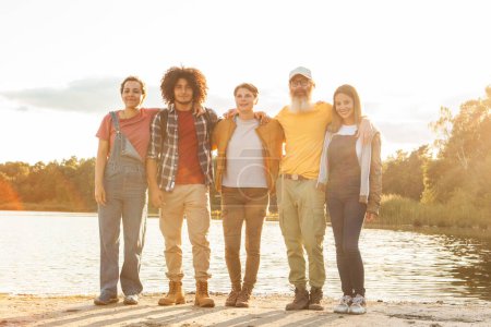 Esta imagen conmovedora captura a un grupo de cinco amigos de pie junto a un lago sereno, bañados por la suave luz dorada del sol poniente. El grupo es un cuadro de unidad en la diversidad