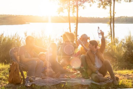 Grupo de diversos adultos y niños, de géneros mixtos y edades, compartiendo una comida junto a un lago, rodeado de naturaleza, en el cálido resplandor del sol de la noche. Familia y amigos disfrutando de un picnic junto al lago en
