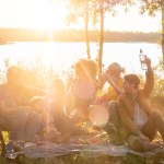 Group of diverse adults and children, mixed genders and ages, sharing a meal by a lake, surrounded by nature, in the warm glow of the evening sun. Family and Friends Enjoying Lakeside Picnic at