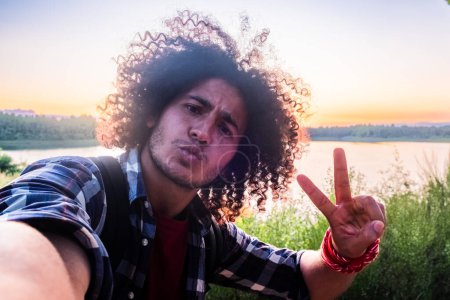 Adulto joven, cabello rizado, dando señal de paz, al aire libre, fondo del atardecer, expresión franca. Curly-Haired Man Making Peace Sign During Sunset Selfie (en inglés). Foto de alta calidad