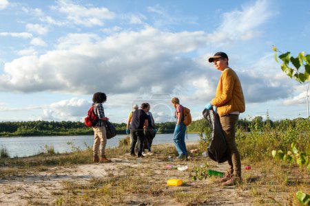 Eine Gruppe verschiedener Freiwilliger sammelt Müll in der Nähe eines Sees mit spärlichen Wolken am Himmel und vermittelt Umweltschutz. Team von Freiwilligen säubert an einem sonnigen Tag das Seegebiet. Hochwertiges Foto