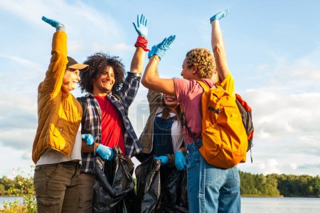 Joyous group of volunteers raising hands in triumph after a lake shore clean-up effort. Ecstatic Volunteers Celebrating a Successful Lake Cleanup. High quality photo