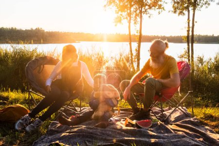 A serene moment captured as a bearded senior man, a young woman, and a child unwind on camping chairs by a lake, surrounded by untouched nature, basking in the golden sunset glow. Family Enjoying