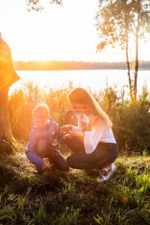 Cette image chaleureuse capture un moment tendre entre une mère et son jeune enfant lors d'un magnifique coucher de soleil au bord du lac. La douce lumière dorée du soleil filtre à travers les arbres, illuminant la scène