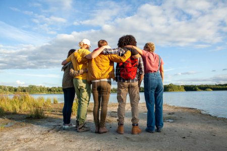 Foto de Abrazada en fila, esta imagen retrospectiva retrata maravillosamente a un grupo de amigos que miran hacia un lago tranquilo, simbolizando un sentido compartido de aventura y contemplación. Sus diversas etnias - Imagen libre de derechos