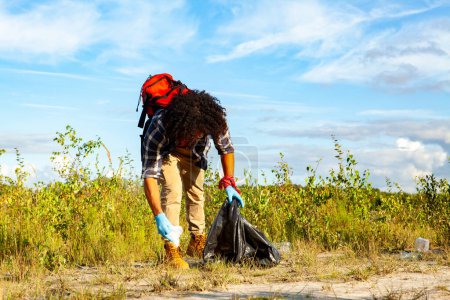 Eine lockige Person mit Handschuhen sammelt in einem Naturgebiet Müll in einer schwarzen Tüte und leistet damit einen Beitrag zum Umweltschutz unter strahlend blauem Himmel. Freiwillige Säuberung von Abfall auf einem Feld auf einem