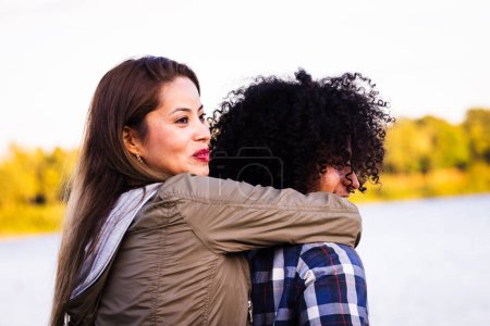 This image captures a woman and a man in a warm embrace, sharing a moment of connection by the lake at dusk. The woman, looking into the distance, has her arm around the man, who has curly hair and is