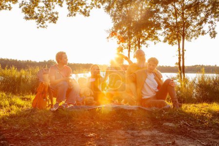 This warm and inviting image captures a serene moment of camaraderie and relaxation among a diverse group of individuals gathered by a lakeside at sunset. The sun, hovering just above the horizon
