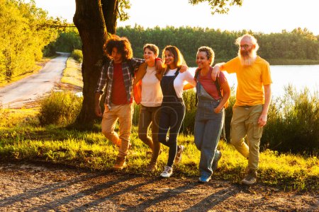 A diverse group of five adults strolls by a tranquil lake, enjoying each others company in the soft light of the setting sun. Diverse Friends Enjoying a Walk by the Lake at Sunset. High quality photo