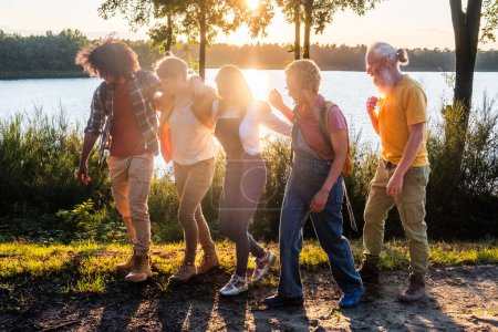 A diverse group of individuals of varying ages and ethnicities enjoying a leisurely walk by a lake during sunset. Multigenerational Friends on Lakeside Stroll at Sunset. High quality photo