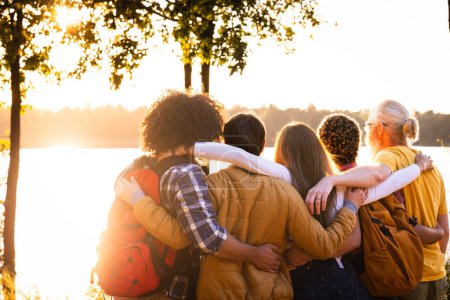 This image is imbued with the warmth of a setting sun as it captures a circle of friends standing by a lake, enveloped in a collective embrace. Each individual is partially facing away from the camera