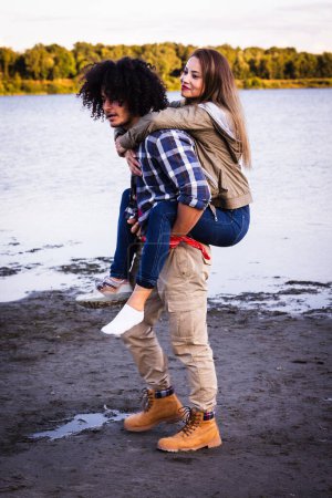 Captured in a candid lakeside setting, this dynamic image features a multiracial couple engaged in a playful piggyback ride. An African American man with curly black hair and a plaid shirt carries a