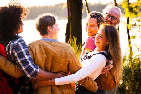 This image exudes warmth and camaraderie as a group of multiethnic friends share a group hug by the lakeside during sunset. Their smiling faces and the closeness of their embrace reflect strong bonds