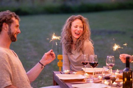 Das Bild fängt eine herzerwärmende Szene ein, in der ein Mann und eine Frau bei einer rustikalen Gartenweinparty in der Dämmerung einen lachenden Moment mit Wunderkerzen genießen. Fröhlicher Abend mit Wunderkerzen im Garten