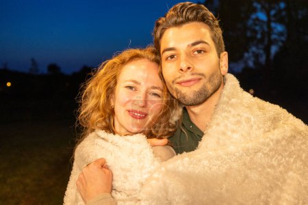This image exudes warmth and affection, capturing a couple wrapped in a fluffy blanket, sharing a close moment with smiles against the twilight of a dusky sky. Warm Embrace: A Couple Enjoying a Cozy