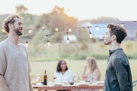 Der sanfte Schein von Lichterketten bereitet den Boden für einen entspannten Abend, wenn zwei Freunde miteinander lachen, während andere im Hintergrund Wein genießen, was die Freude an geselligen Zusammenkünften im Freien verkörpert. Abend