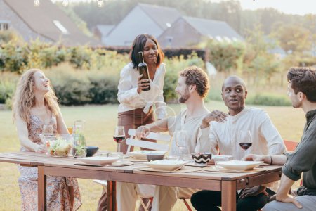 Un groupe d'amis profite d'un moment de rire et de vin dans un jardin ensoleillé, créant une atmosphère de loisirs et de plaisir lors d'une dégustation de vin d'été décontractée. Dégustation de vin d'été avec des amis