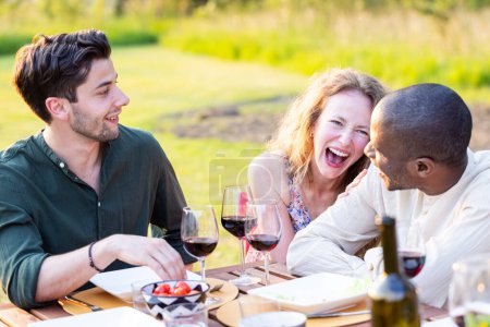 Inmitten des goldenen Stundenglühens lacht ein befreundetes Trio herzhaft über einen Tisch mit Wein und Häppchen und genießt die Freude an einem entspannten Dinner im Freien. Lachen und Wein: Fröhliches Al Fresco