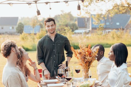 In the soft radiance of the setting sun, a man stands with a gentle smile as he engages in lighthearted conversation with friends seated around a rustic table adorned with wine glasses and lush
