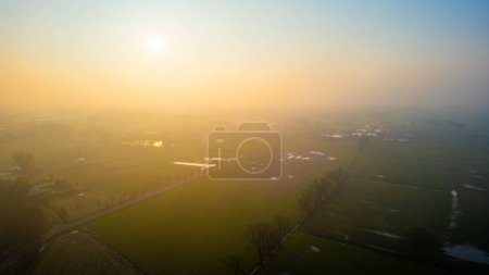 Sunrise envelops the countryside in a misty glow, revealing scattered pools amid the fields in this pastoral scene. Misty Morning Glow Over Rural Landscape with Patches of Water. High quality photo