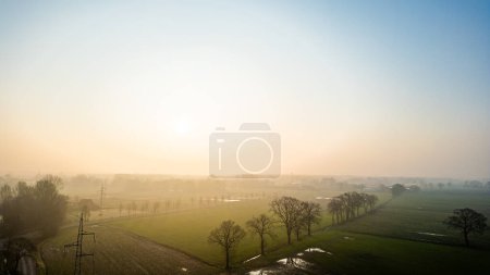 The suns gentle rise diffuses through mist, casting a hazy glow over a quiet rural expanse dotted with trees. Soft Dawn Light Over a Misty Rural Landscape with Scattered Trees. High quality photo