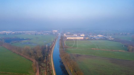 A hazy morning scene unfolds, revealing the juxtaposition of agricultural fields and industrial development from above. Elevated View of Misty Canal Dividing Rural Farmland and Industrial Estate. High