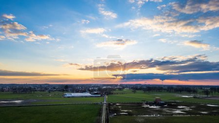 As the sun sets, its rays peek through cloud cover, casting a dramatic light over expansive farmland and facilities below. Sunset Sky Over Farmland: A Majestic Display of Clouds and Light. High