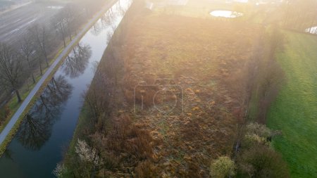 Sunrise bathes a serene canal and path in a warm, hazy light, highlighting the tranquility of the natural landscape. Misty Aerial View of a Lush Canal Path at Sunrise. High quality photo