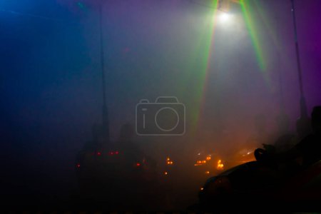 Dieses Bild zeigt die lebhafte und verschwommene Atmosphäre eines Rummelplatzes mit Autoscooter inmitten eines dichten Nebeleffekts. Die pulsierenden Lichtstrahlen der Overhead Ride Struktur durchschneiden die