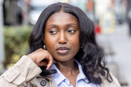 La fotografía muestra a una joven africana en pensamiento contemplativo en medio de un telón de fondo urbano. Ella está vestida con elegancia en una gabardina sobre una camisa de rayas azules, personificando una mezcla de profesionales