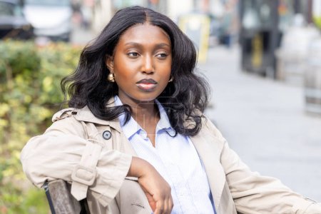 A young black woman is pictured seated outdoors with a contemplative gaze. She wears a smart casual outfit consisting of a light blue striped shirt and a classic beige trench coat. Her long black hair