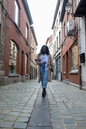 This image features a stylish woman walking down a cobblestone street in a historic European neighborhood. She is fashionably dressed in a blue collared shirt, high-waisted jeans, a beige trench coat