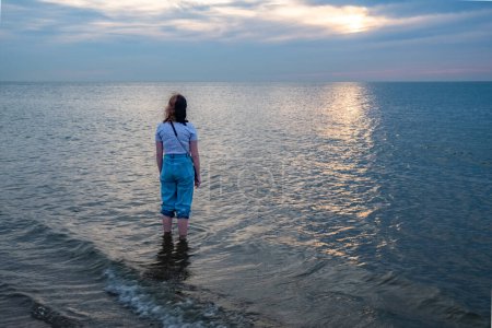 Dieses ergreifende Bild fängt eine einsame Person ein, die im Meer steht und auf den fernen Horizont blickt, wo die untergehende Sonne einen silbrigen Weg über das Wasser wirft. Die Personen lässig gekleidet