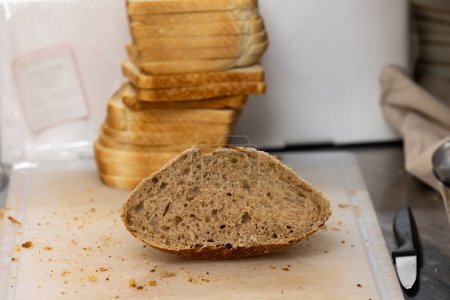 The image captures a half-slice of whole grain bread resting on a wooden cutting board with crumbs around it, suggesting the slicing process has just occurred. In the background, stacked slices of