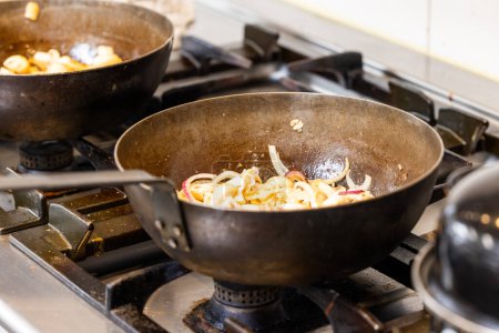 Cette image donne un aperçu des coulisses d'une cuisine commerciale avec deux woks sur des cuisinières à gaz, l'un avec des oignons mi-saute et l'autre avec des champignons. Les woks semblent bien assaisonnés et utilisés, ce qui est
