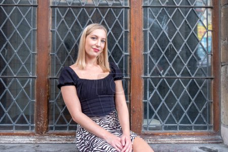 A charming young woman with blonde hair offers a gentle smile while sitting on the ledge of an aged window with leaded glass. Her relaxed pose and the soft light reflecting on her face create an