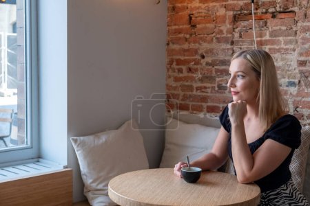 In a serene cafe setting, a young blonde woman is portrayed enjoying a moment of solitude. She sits at a wooden table, gazing thoughtfully out of a large window, with soft daylight illuminating her
